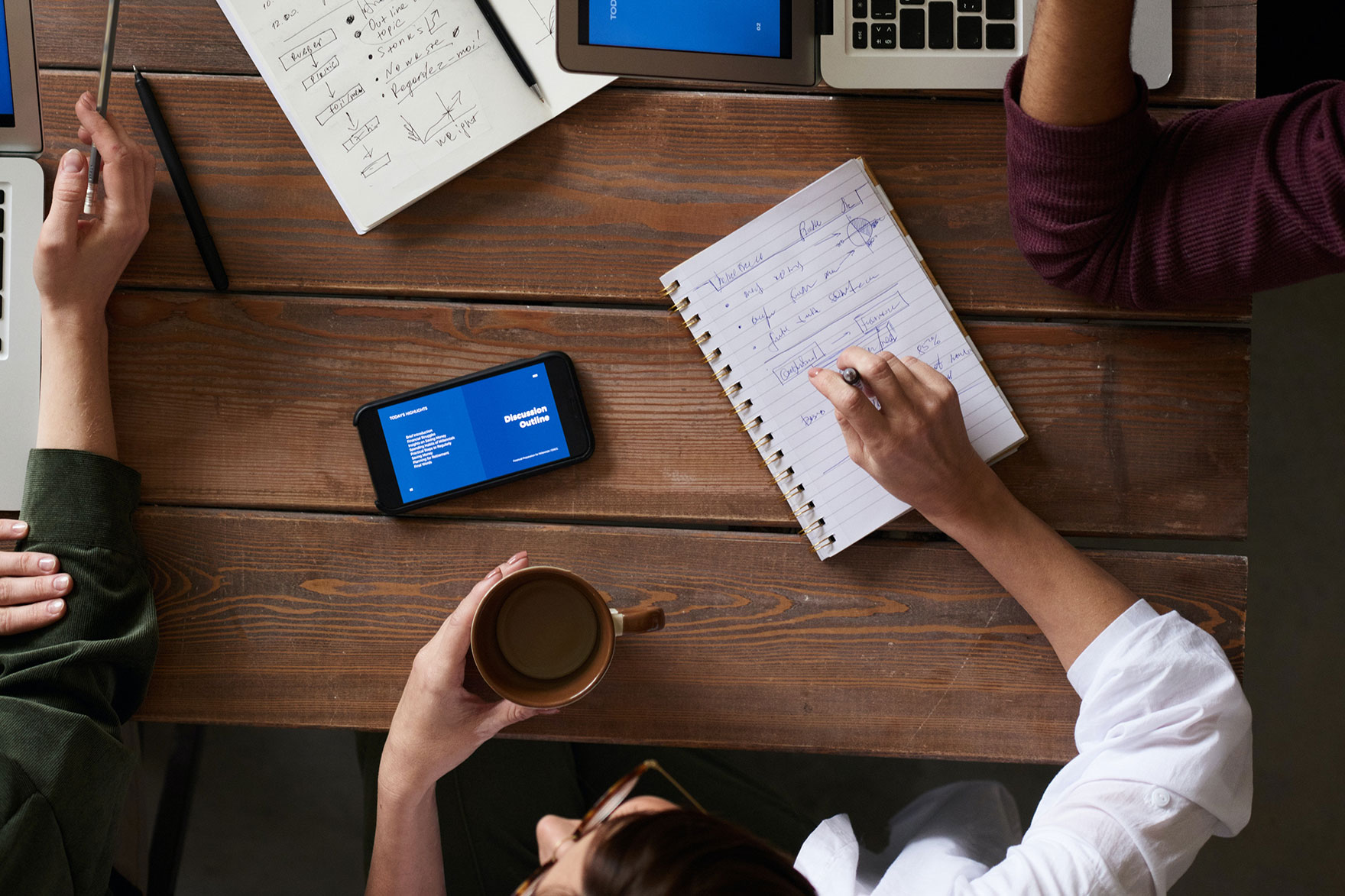 A group of four people sitting at a table discussing strategy with computers and notebooks in front of them shown from the top perspective looking down on the table